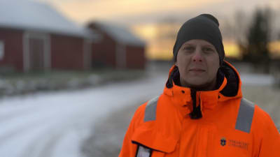 En man med orange arbetsjacka står ute i ett snöigt landskap med en gammal ladugård i bakgrunden. 