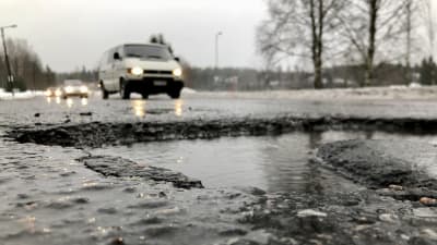 En stor, vattenfylld grop i beläggningen på en asfalterad väg där flera bilar kör.