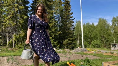 Maria Haglund står bakom sin pallkrage med sallad som hon odlat. Hon har en klänning på sig och håller i en vattenkanna.