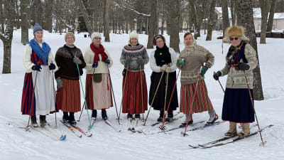 Sju damer iklädda folkdräkt och skidor poserar i Kapellparken i Lovisa.