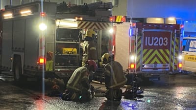 Tre personer i brandkårsutrustning gör nåt med utrustning framför en brandbil.