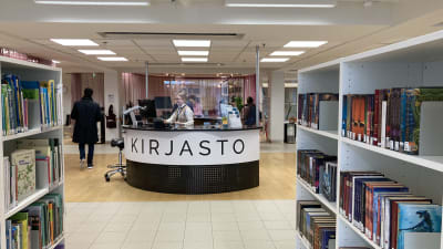 KIrjahyllyjä ja palvelutiski, jossa lukee KIRJASTO. Taustalla asiakkaita ja kirjastotyäntekijä.
