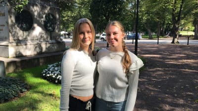 Två unga kvinnor med vita tröjor och blont hår står i park.