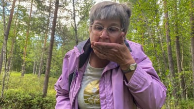 En kvinna med grått hår och lila jacka i en skog. Hon häller blåbär från handen i sin mun.