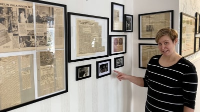 En kvinna står bredvid en vägg med många inramade fotografier och tidningsurklipp. Hon pekar mot en bild.