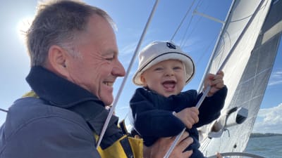 Tapio Lehtinen seisoo tuulen kallistamassa purjeveneessä tyttärenpoika Emil sylissään. Molemmilla on iloinen ilme.