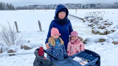 En kvinna och två små flickor står och poserar för kameran intill en barnvagn med en sovande baby. Bakom dem syns is på havet.