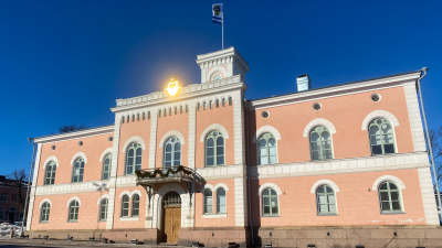 Ett ljusrött och vitt ståtligt hus, Lovisa rådhus. Solen reflekterar i stadens vapensköld högt upp ovanför ingången.