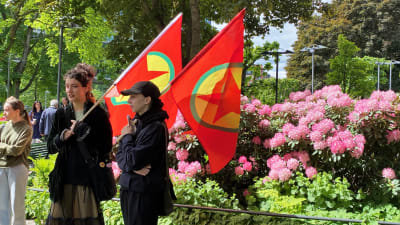 Två kvinnor håller i PKK-flaggor på en demonstration utomhus vid en park.