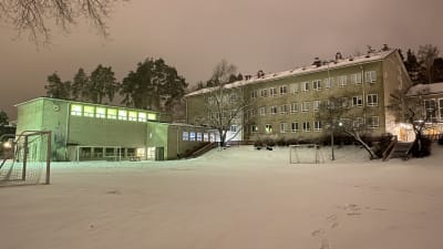 En 1950-talsskola i Karis, grå, rappad, skymning. Lite snö på marken. Belysning i en del av byggnaden. 