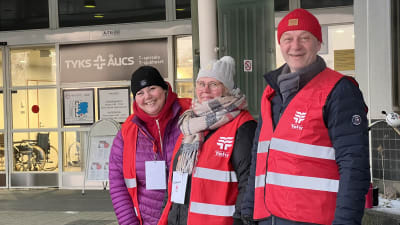 Tre personer ler utanför sjukhusets dörrar. De har på sig röda strejkvästar. 