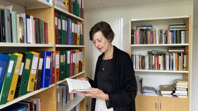 En mörkhårig kvinna som håller i en bok framför en bokhylla.