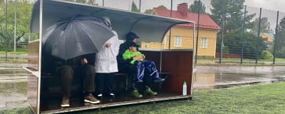 Flera personer med regnjacka och paraply står under skydd vid en fotbollsplan.