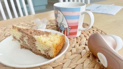 En vibrator ligger på köksbordet intill en kaffekopp, en napp och en tallrik med en tårtbit. 