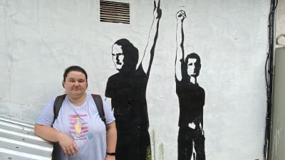 Kvinna i grå t-skjorta står vid en väggmålning föreställande två oppositionella musiker från Belarus. Den ena musikern i målningen visar segertecknet, den andra håller upp en knuten näve.