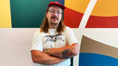 En glasögonprydd Kent Ketomäki iklädd t-skjorta och keps står med armarna i kors framför en färggrann vägg.