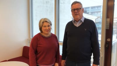 En kvinna och en man står bredvid varandra framför en soffa och ett fönster. Kvinnan har en vinröd tröja på sig och axellångt hår, mannen har glasögon 