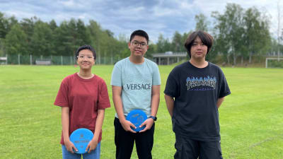 Tre ungdomar står framför en idrottsplan. Två av dem håller i varsin frisbee.