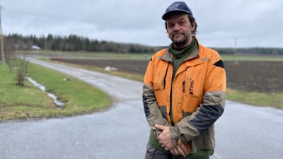 En man i orange jacka och blå keps står framför en åker. 