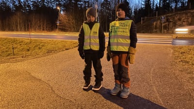 Två barn i vinterkläder och gula reflexvästar står framför ett övergångsställe över en landsväg.