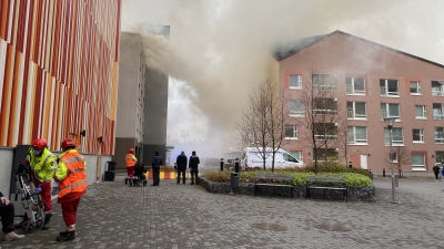 Rykande höghus, människor och brandmän på gatan.