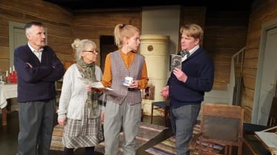 Fyra personer på en teaterscen som föreställer en stockstuga med inredning från 1940-talet.