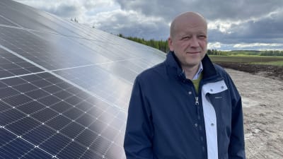 En man med mörkblå jacka står framför en stor solpanel på en åker. 