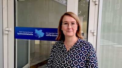 En kvinna i glasögon utanför en dörr till Egentliga Finlands välfärdsområdes lokaler. 