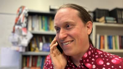 Man i röd skjorta står framför en bokhylla och talar i mobiltelefon