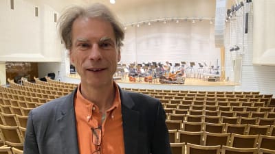 Eckhard Kahle, en man med grånande hår och grånande buskiga ögonbrun, står i en orange skjorta med en orkester på en scen i bakgrunden.