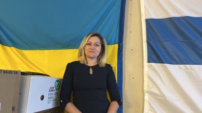 Kvinna i mörkblått ler mot kameran. I bakgrunden syns en ukrainsk och finsk flagga.