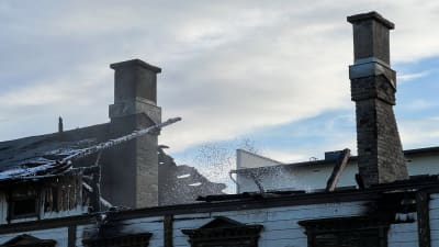 Taket på det vita trähuset har rasat. Räddningstjänsten sprutar in vatten i öppningen i taket. Det finns en förkolnad tunna i stående.