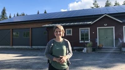 Kvinna ler mot kameran. Hon står framför ett rött hus med solpaneler på taket.
