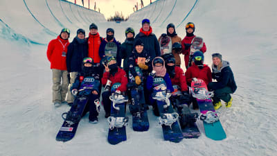 En grupp leende människor står tillsammans med sina snowboards i en snötäckt "halfpipe", alltså en slags bana för snowboarding. 