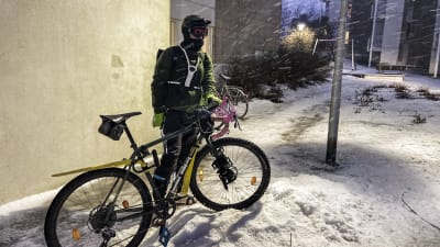 En person står intill en cykel, med vintercyklingsutrustning. Det snöar.