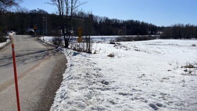 En asfalterad väg till en plankorsning. Till höger syns banan och snöiga åkrar. Vårvintersol och snö.