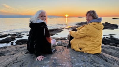 Maarit Tastula ja Outi Airola istuvat auringonlaskussa meren rannalla kalliolla.