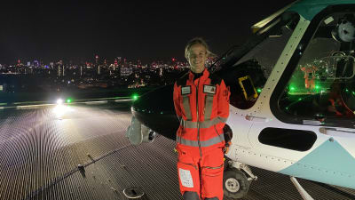 Arbetsklädd kvinna ler och ser in i kameran, står framför en ambulanshelikopter. Det är kväll eller natt och i bakgrunden syns silhuetten av centrala London.