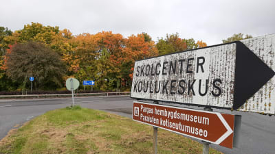 En vit vägskylt med texten Skolcenter Koulukeskus, och en brun skylt med texten Pargas hembygdsmuseum, i en vägkorsning med höstruska i träden i bakgrunden.