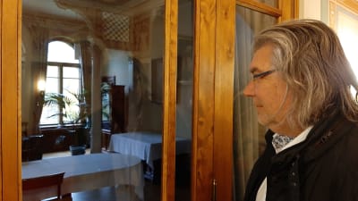 Mikael Lindfelt, en man med glasögon och axellångt grånande hår, tittar in genom ett fönster till en sal i ett 1800-talshus.