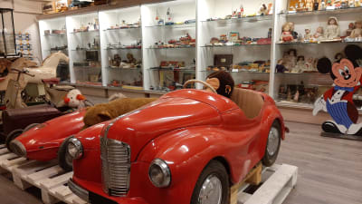 En stor röd trampbil mitt på golvet bland en stor mängd leksaker i ett leksaksmuseum.