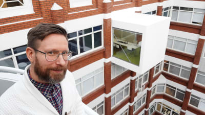 Nikolas Salomaa, en man med glasögon, mustasch och skägg, står på en balkong mellan höga hus.