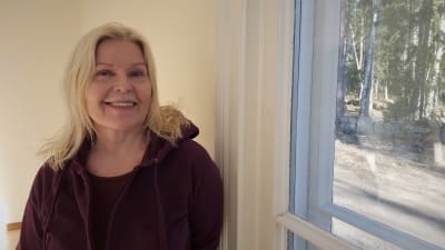 Blond kvinna poserar vid ett fönster.