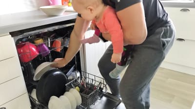 En pappa med sin baby i bärsele fyller diskmaskinen.