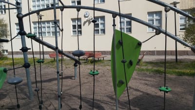 Ljus skolbyggnad med klocka på väggen och klätterställning i förgrunden.