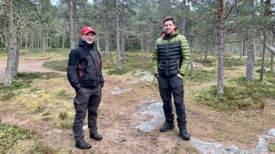 Två män står i skogen med friluftskläder. 