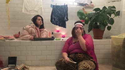 Två tjejer sitter i ett sunkigt badrum. Den ena sitter i badkaret med en dunjacka på och en cigarett i mungipan, medan andra sitter med solglasögon och keps på golvet.