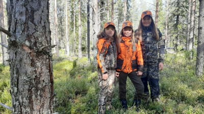 Tre flickor i jaktkläder i skogen.