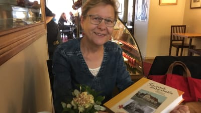 Riitta Lehtonen sitter på ett café och håller boken Terveisiä sairaalasta i sin hand.