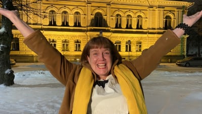En glad kvinna i vinterkläder lyfter händerna i en segergest. Bakom henne syns Borgå stadshus.